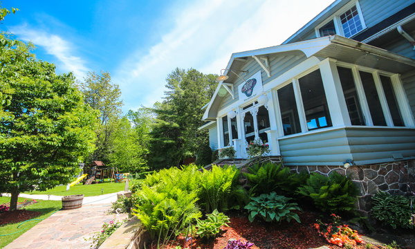 A grey sided building with white trim is pictured in front of a lush garden with ferns and trees